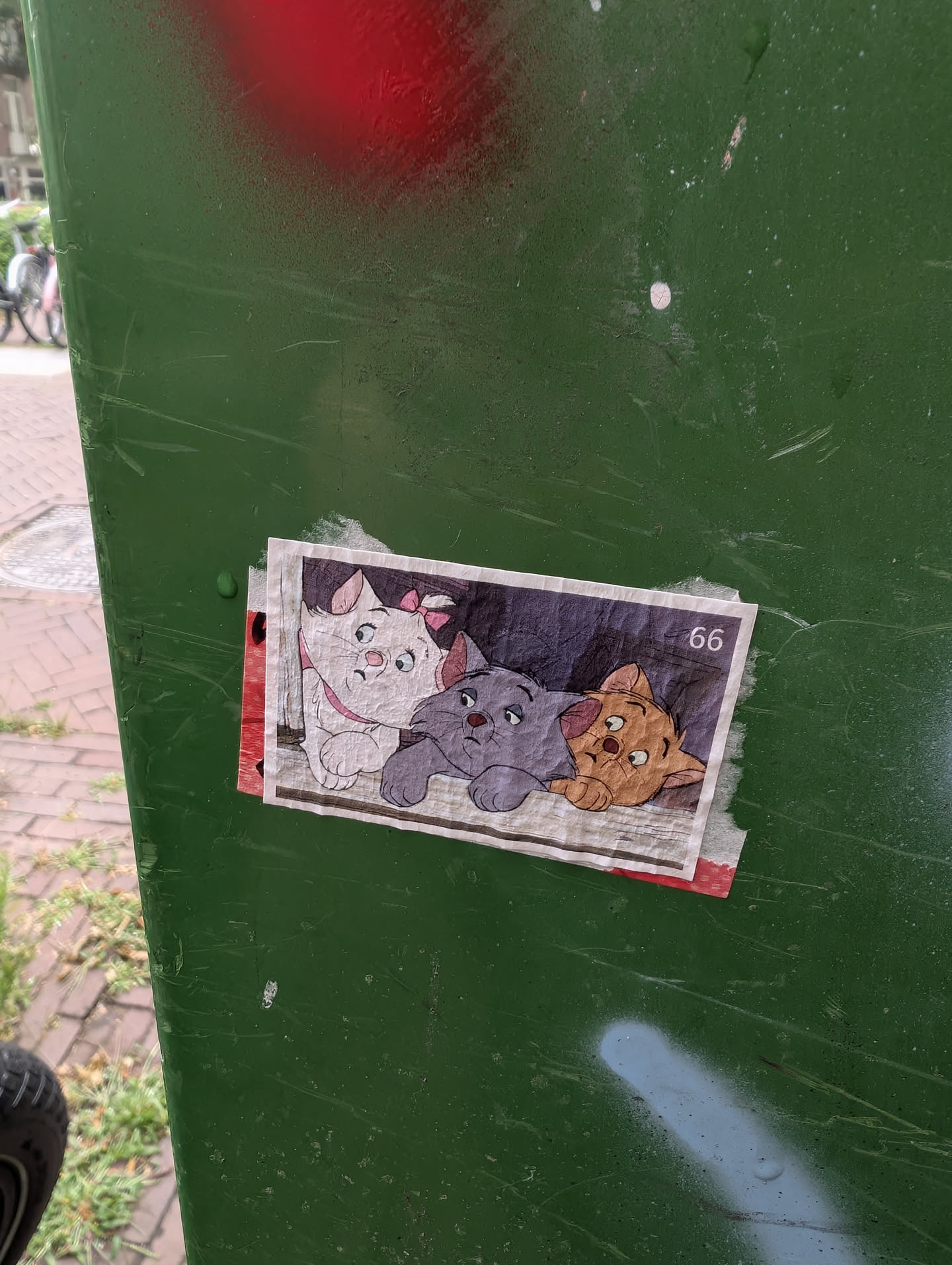 The sticker features three cartoon cats with distinct colors and expressions, positioned side-by-side. The leftmost cat is white with a pink bow and collar. The middle cat is grey with a grey collar. The rightmost cat is orange-brown with a red bow. They are all depicted with large eyes and a slightly melancholic expression. The background of the sticker is a faded greyish-white, with what appears to be printed lines. In the upper right corner of the sticker, the number '66' is visible in black. The sticker has a slightly worn appearance and is attached to a green surface with some peeling edges, suggesting it has been adhered for some time.