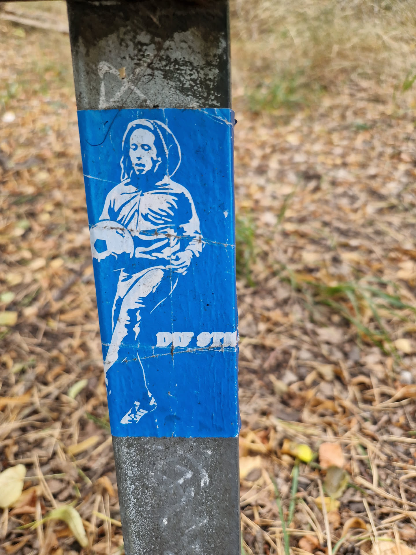 A blue sticker affixed to a metal post depicts a white stencil of a person, possibly a soccer player, holding a ball. The style suggests street art or graffiti.  The text 'DU STIM' is partially visible at the bottom. The sticker appears slightly damaged or worn.