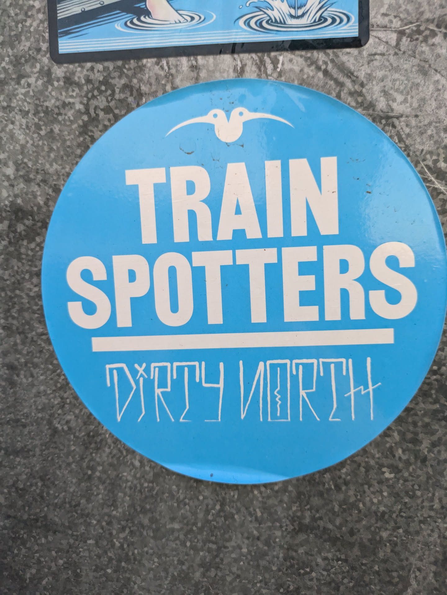 A round blue sticker with the text "TRAIN SPOTTERS" in white uppercase letters. Underneath, "DIRTY NORTH" is written in a stylized font.  A small bird-like symbol is at the top.