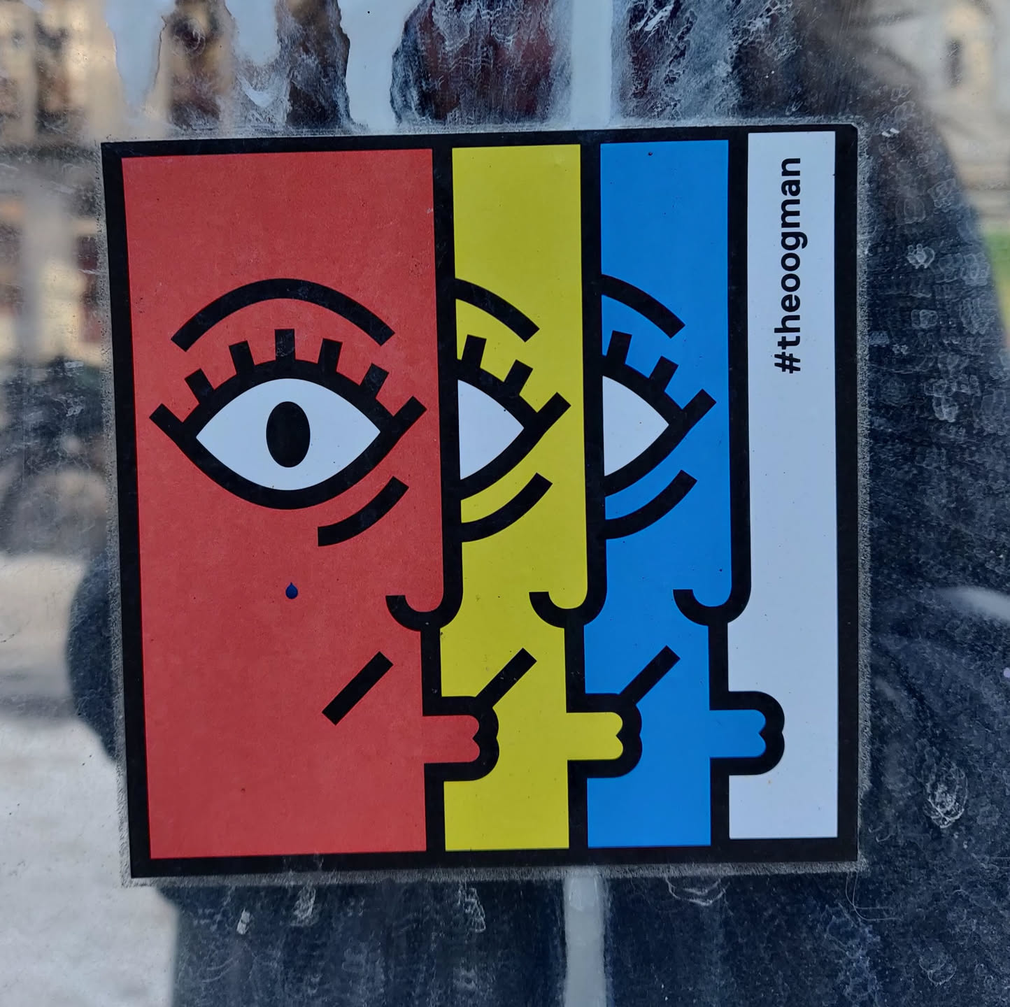 A sticker with a geometric design featuring three stylized faces, each a different color: red, yellow, and blue. The faces are connected, sharing features, and are depicted in a simple, bold style.  The hashtag #theoogman is visible on the sticker.