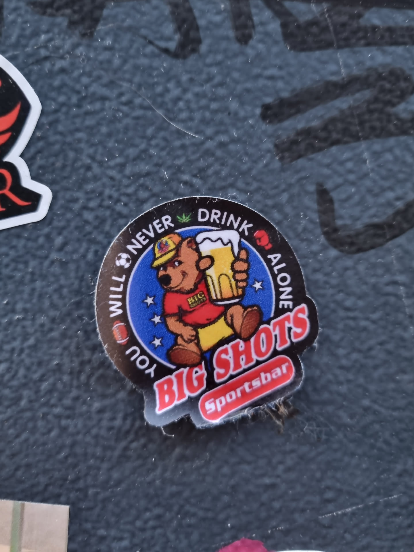 A round sticker featuring a cartoon bear holding a beer mug. The text around the bear says "You will never drink alone" with some icons and the text "BIG SHOTS Sportsbar" at the bottom. 