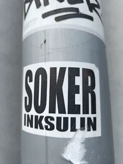 A black and white sticker with the words "SOKER" stacked on top of "INKSULIN" in a bold, sans-serif font. The sticker shows some wear and tear from being outside. It's been stuck on a metallic grey pole.