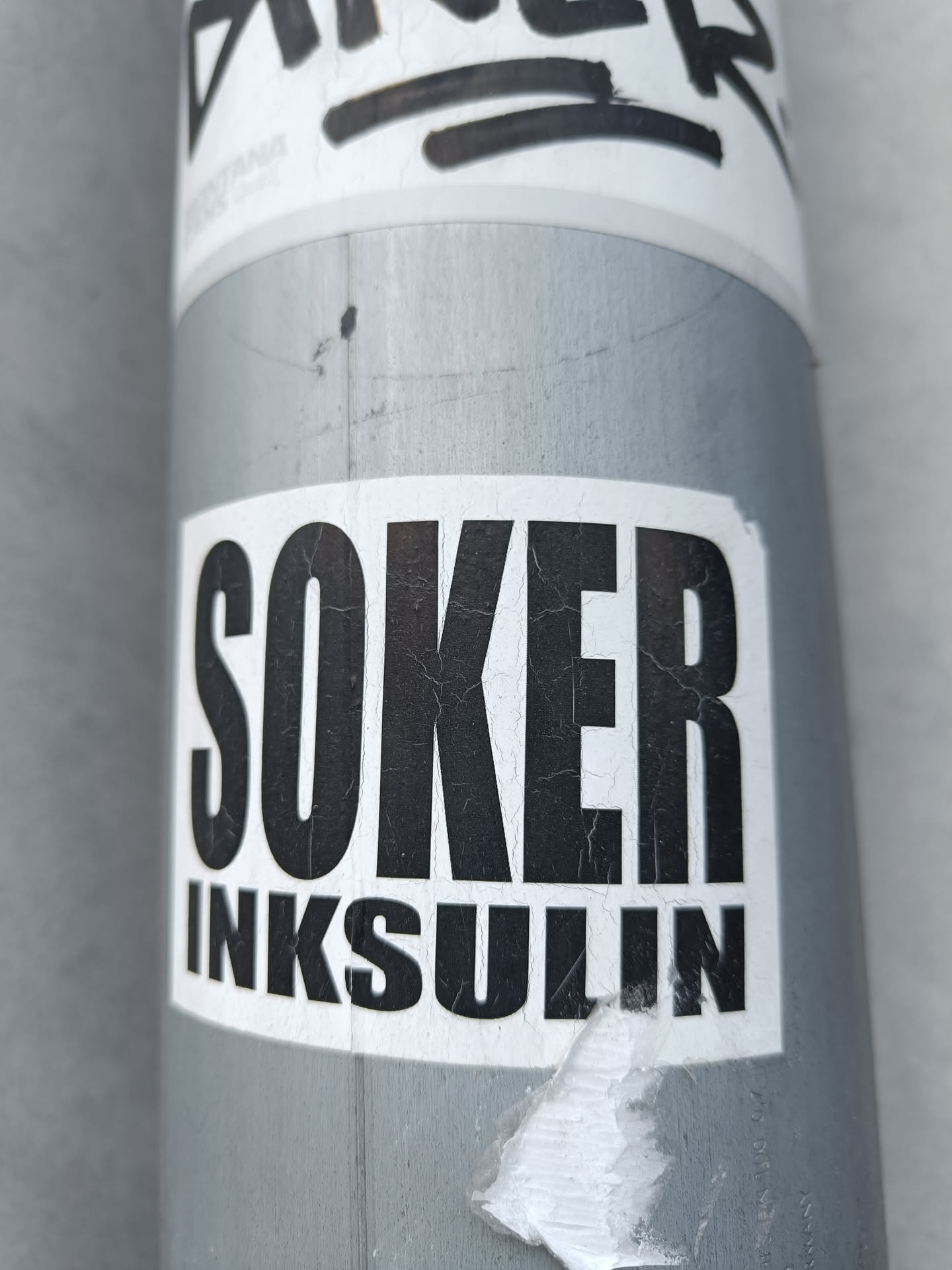A black and white sticker with the words "SOKER" stacked on top of "INKSULIN" in a bold, sans-serif font. The sticker shows some wear and tear from being outside. It's been stuck on a metallic grey pole.
