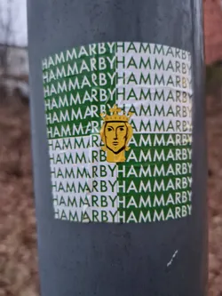 Close-up of a sticker featuring a golden crowned figure on a background with repeating "Hammarby" text in green and white. The sticker is affixed to a gray pole.