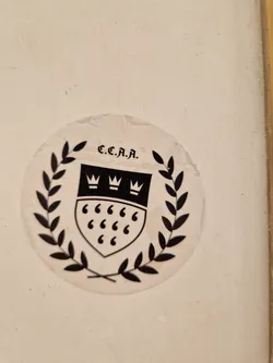 Close-up of a round sticker featuring a black and white design. The sticker shows a shield with three crowns on top and quotation marks inside, surrounded by a laurel wreath. The text "C.C.J.A." is visible above the shield. The sticker appears to be affixed to a light beige surface.