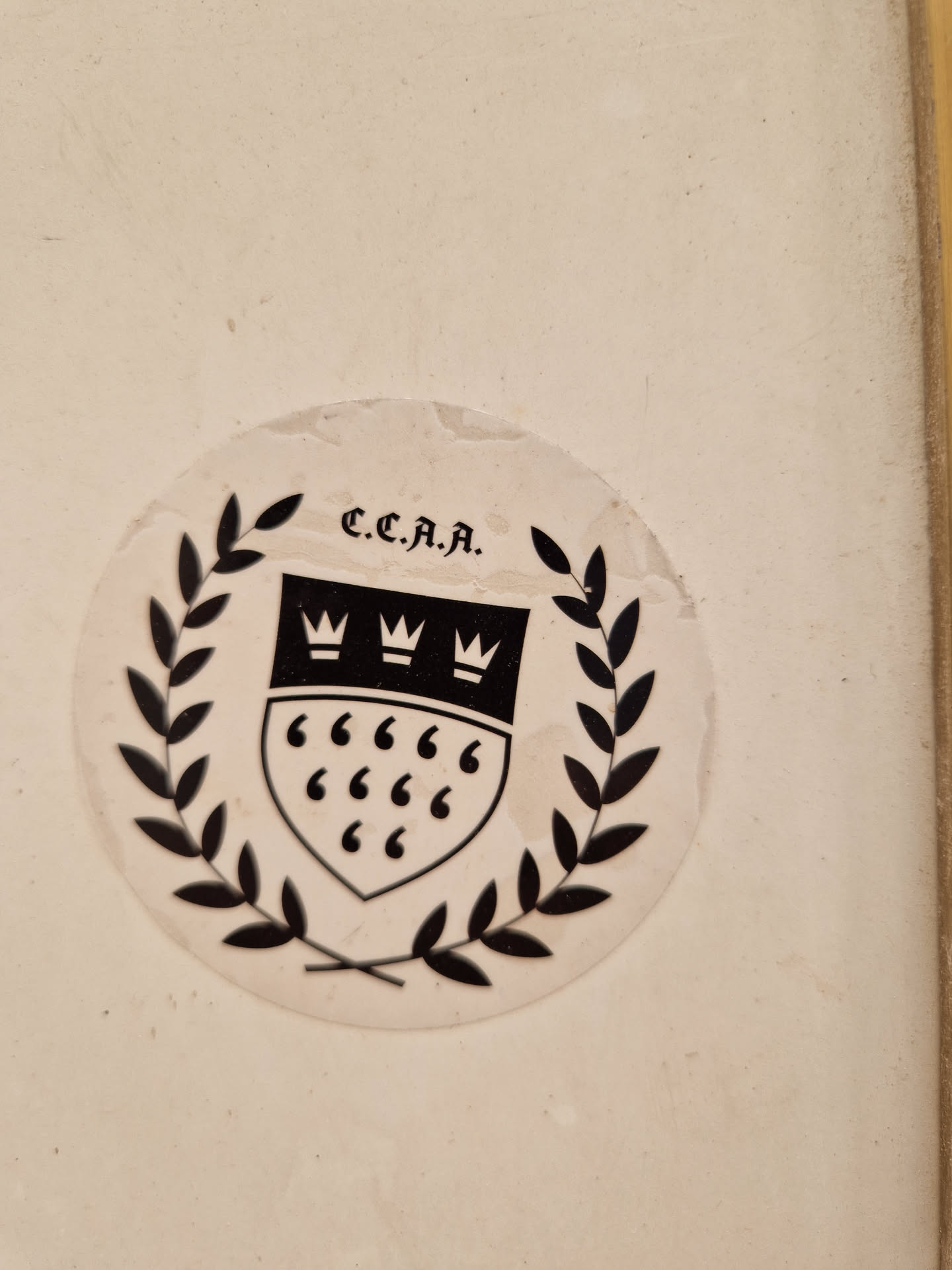 Close-up of a round sticker featuring a black and white design. The sticker shows a shield with three crowns on top and quotation marks inside, surrounded by a laurel wreath. The text "C.C.J.A." is visible above the shield. The sticker appears to be affixed to a light beige surface.
