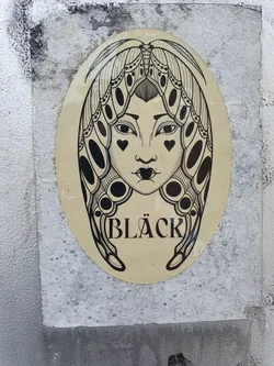 Oval sticker featuring a monochrome illustration of a woman's face framed by moth wings. The word "BLACK" is written at the bottom. The style is reminiscent of tattoo art.