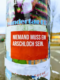 A red sticker with a German phrase on it, pasted on a pole covered in other posters. The phrase promotes kindness and discourages negativity.