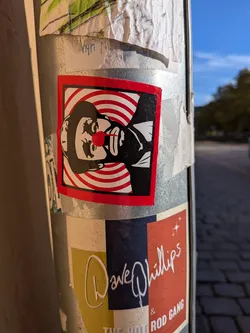 The sticker is square-shaped with a thick red outer border. In the center, set against an alternating red-and-white concentric circle pattern, is a high-contrast black and white stencil portrait of a beret-wearing man (reminiscent of Che Guevara). A bright red clown nose, extending downwards slightly like a teardrop or drip, covers his nose, giving the serious portrait a satirical, humorous twist.