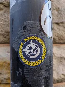 A round sticker of the sports club SK Vöest is on a black metallic pole. The sticker is framed with a golden laurel. At the top center is a logo with two crossing bars and the words "SK VÖEST".