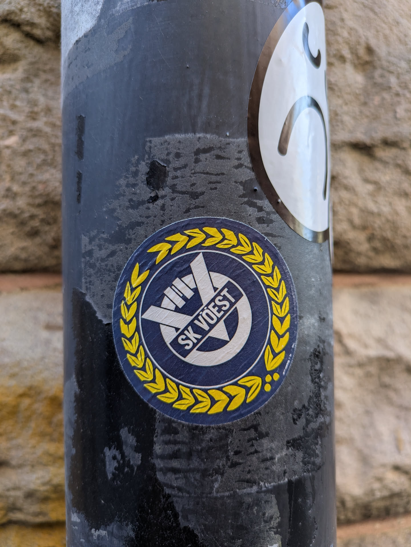 A round sticker of the sports club SK Vöest is on a black metallic pole. The sticker is framed with a golden laurel. At the top center is a logo with two crossing bars and the words "SK VÖEST".