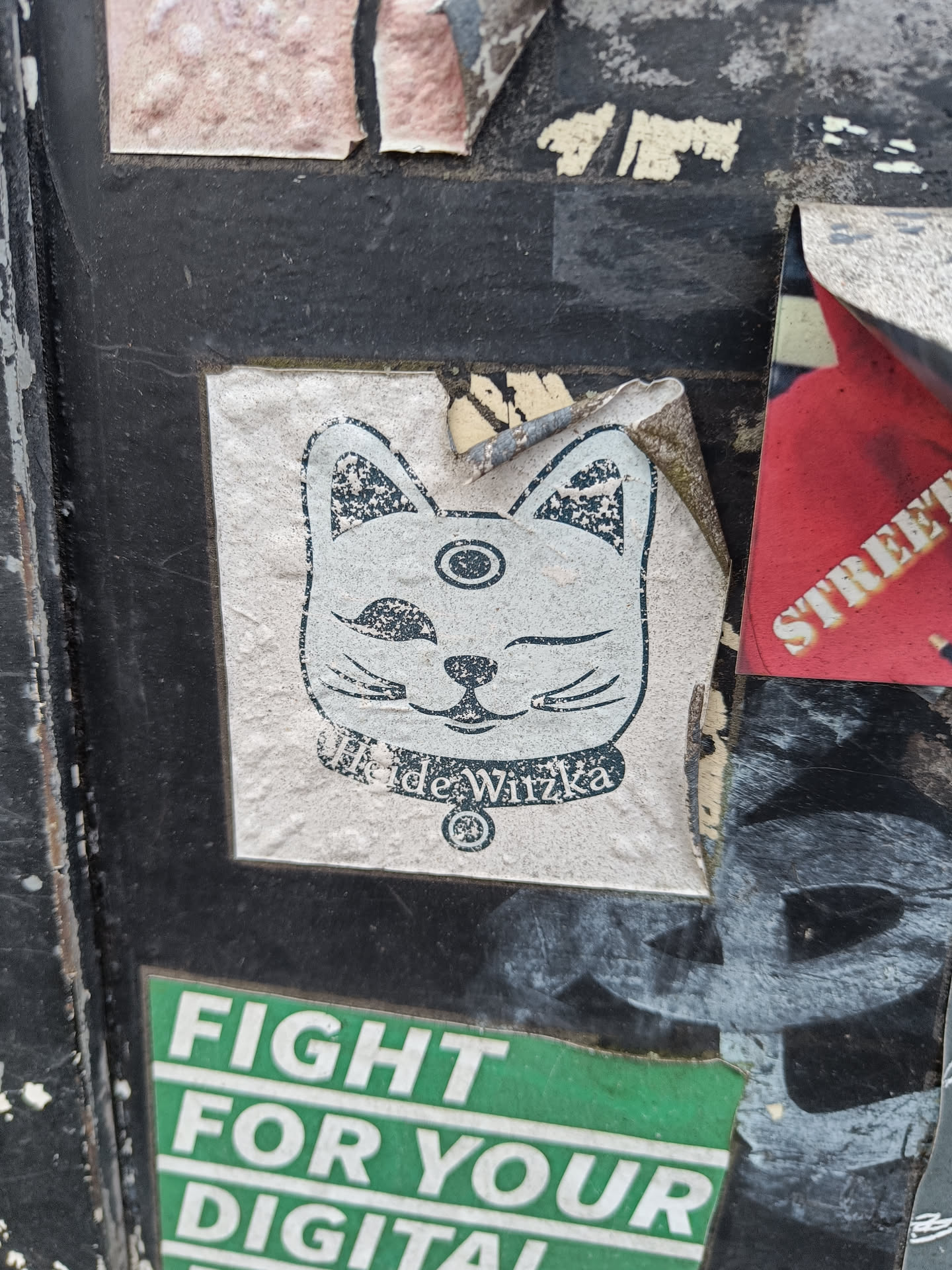 Close-up of a sticker featuring a stylized Maneki-neko (beckoning cat) with a third eye.  The artist's name, Ilde Wirzka, is visible at the bottom. The sticker is affixed to a surface covered in other stickers and graffiti.