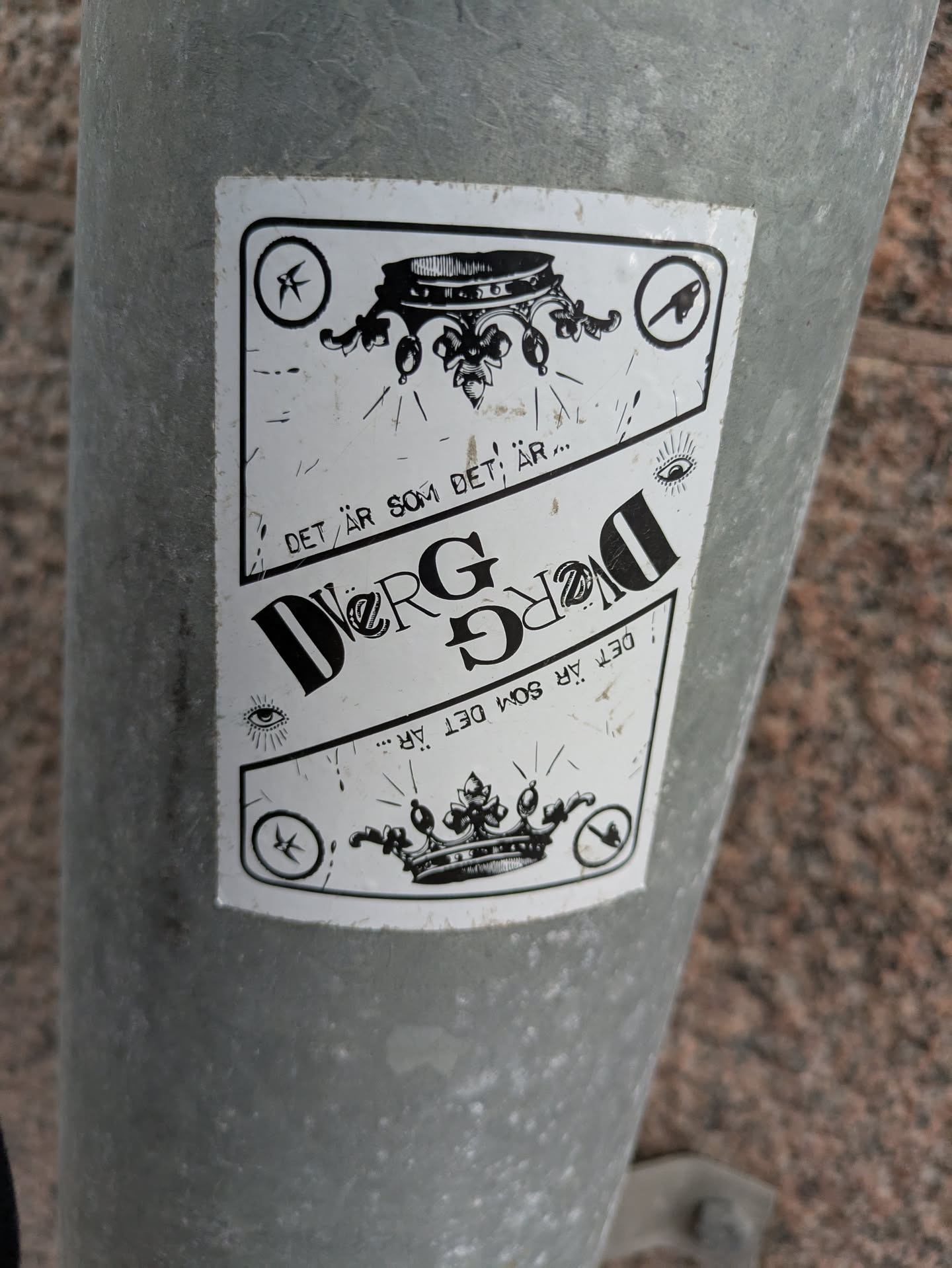 This sticker shows a monochrome design with a crown as a central element. The text "DET ÄR SOM DET ÄR..." is repeated vertically on both sides of the crown. There are also additional symbols, likely adding to the visual message or brand.