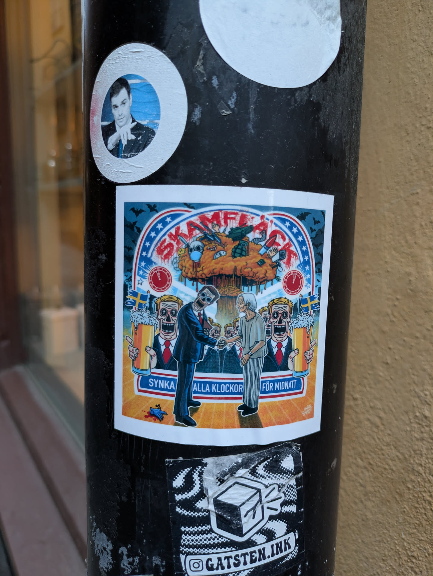 A sticker with a dark humor illustration depicting a meeting between two skeletal figures representing political ideologies, with a mushroom cloud in the background. The text 'Skamflck' is prominent, along with 'Synka alla klockor för midnatt' (Synchronize all clocks for midnight). The style is reminiscent of political satire and dark art.