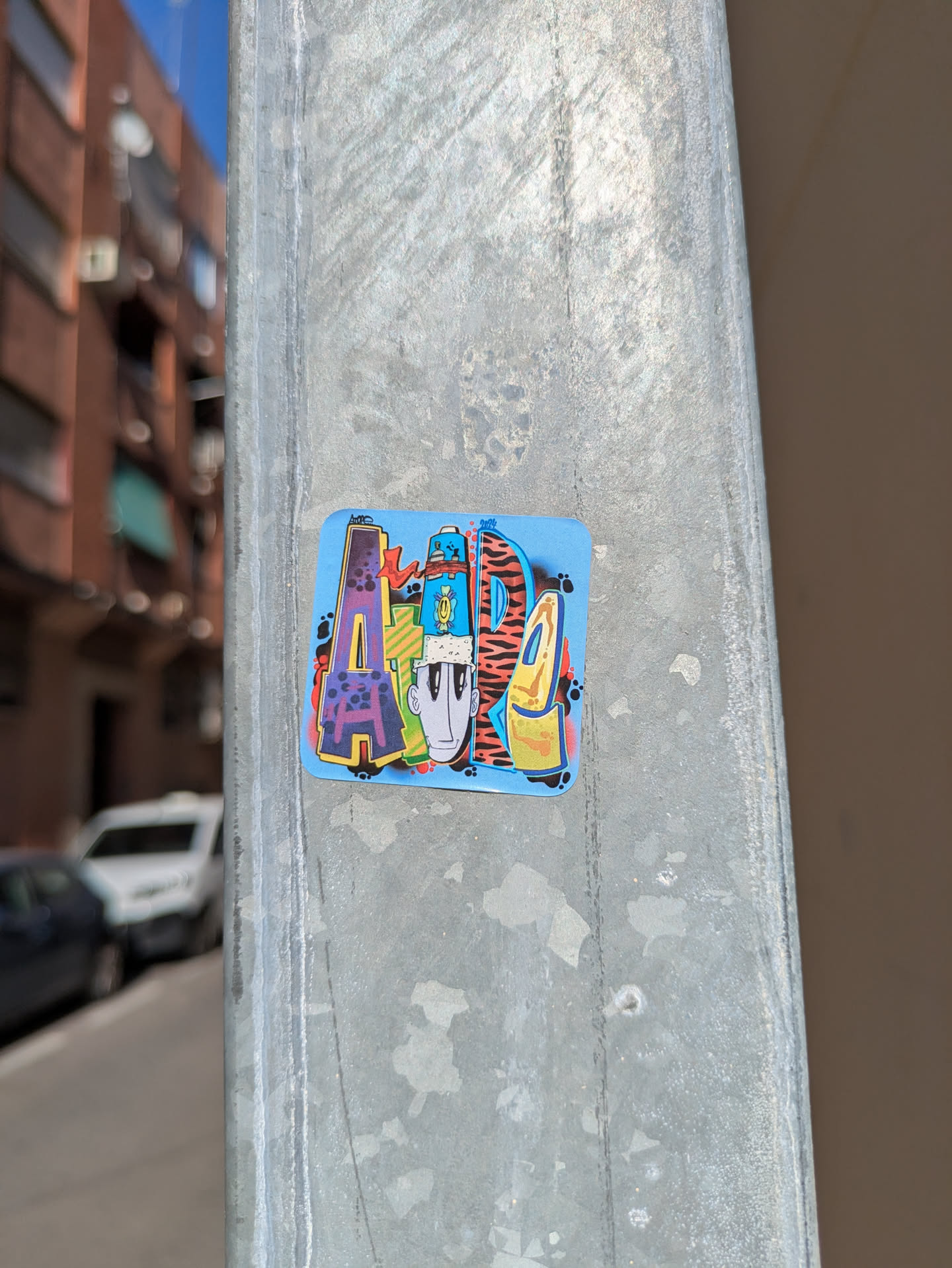 A square sticker with rounded corners, adhered to a metal pole, features an artistic, graffiti-style design. The sticker has a light blue background, with the stylized letters 'ARDI' prominently displayed. The letters are filled with different textures and colors, with a character face integrated within the design. The sticker looks like its applied in an urban environment.