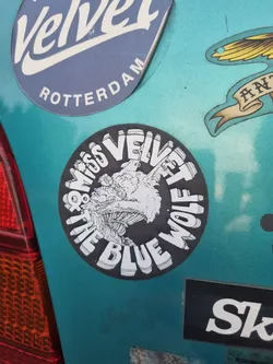 A round sticker featuring a stylized wolf design and the text "Miss Velvet & the Blue Wolf" in a circular pattern. The sticker has a black background with white text and graphic.