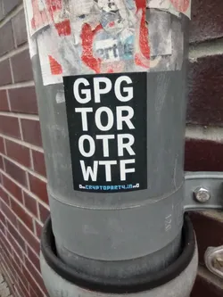A sticker with the text "GPG TOR OTR WTF" and the website address "On.CryptoParty4.io" is affixed to a pipe. The sticker is black with white text and has a minimalist design. The background shows a brick wall.