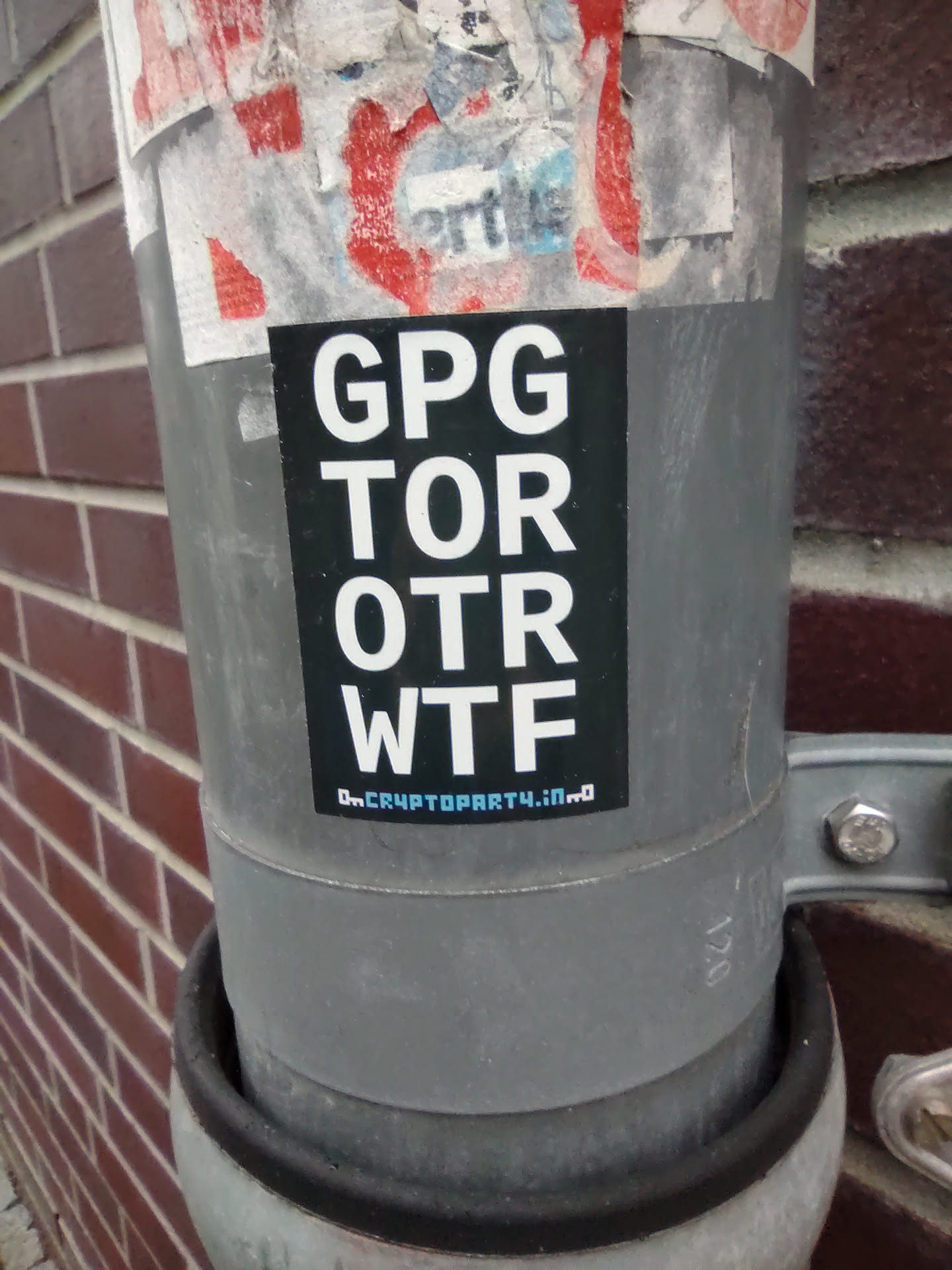 A sticker with the text "GPG TOR OTR WTF" and the website address "On.CryptoParty4.io" is affixed to a pipe. The sticker is black with white text and has a minimalist design. The background shows a brick wall.