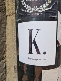 A minimalist sticker featuring a large capital K and a period, with a website address below. The sticker is affixed to a dark-colored pole.