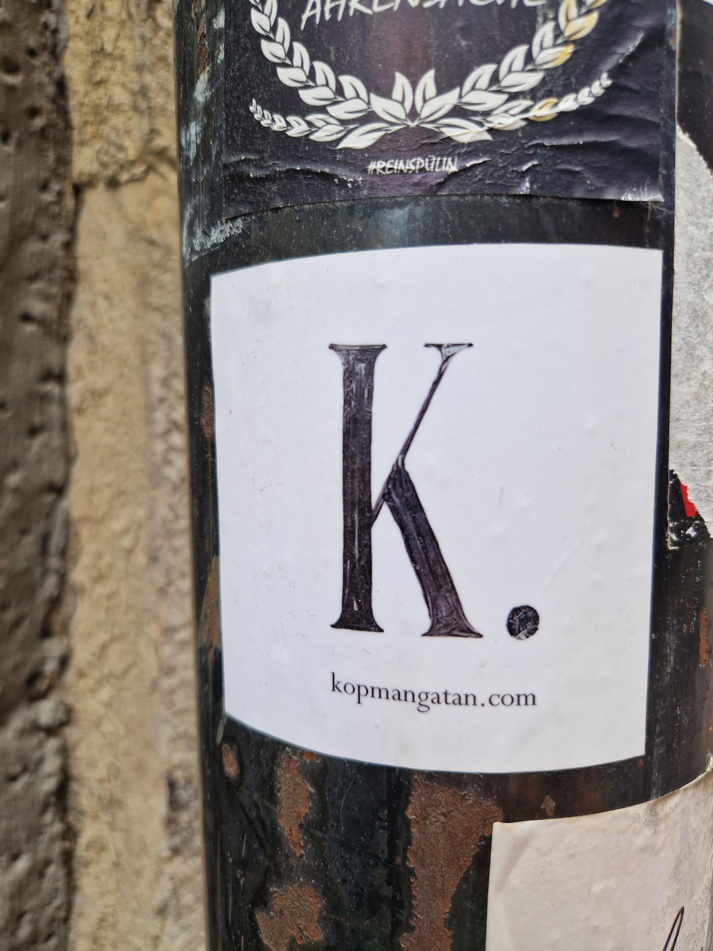 A minimalist sticker featuring a large capital K and a period, with a website address below. The sticker is affixed to a dark-colored pole.