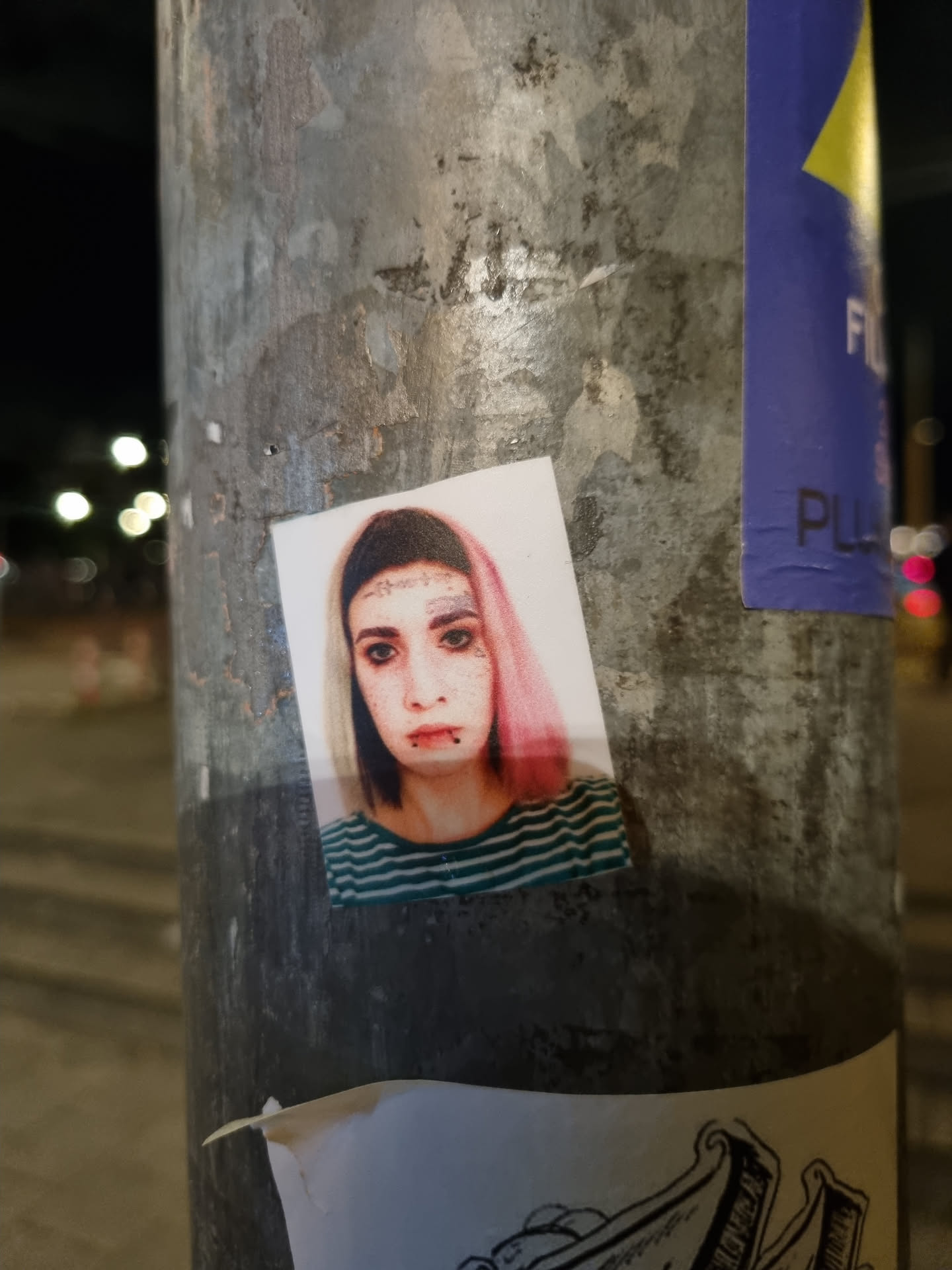 A sticker featuring a close-up portrait of a young woman with pink hair, facial piercings, and a striped shirt. The sticker is affixed to a weathered metal pole outdoors.