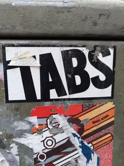 A black and white sticker with the word "TABS" in a bold, sans-serif font. The sticker is affixed to a weathered gray surface.  There are other, partially torn stickers visible underneath.