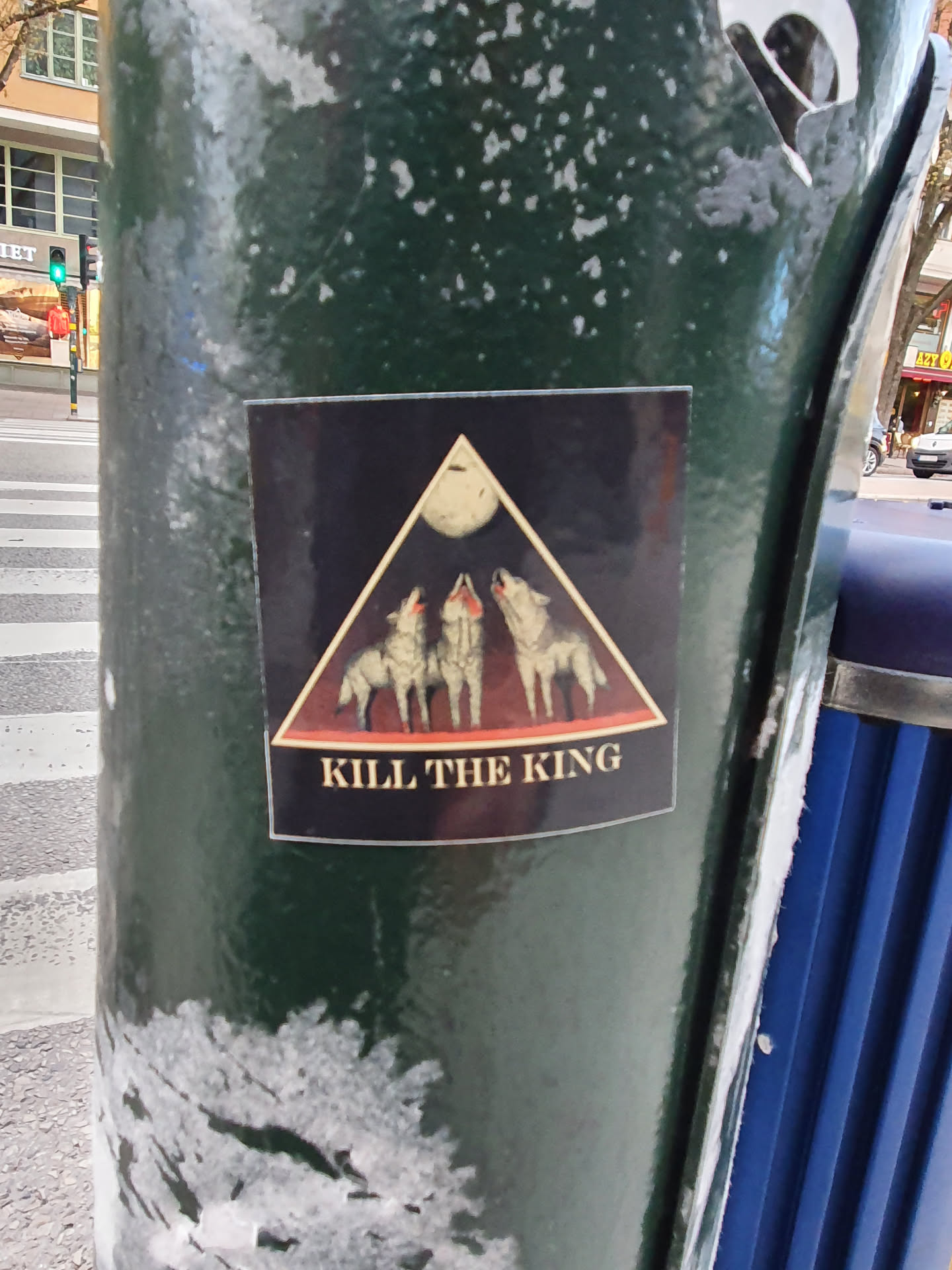 A sticker featuring three wolves howling at the moon within a triangle. The words "KILL THE KING" are written below the image. The sticker has a dark background and is placed on a dark green pole.