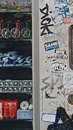 The image features a wall covered in various stickers, including one that reads "SAME", "ONTUE", "ABL SUBS AT LOS ANGELES". There's also graffiti and other markings adding to the wall's textured appearance.