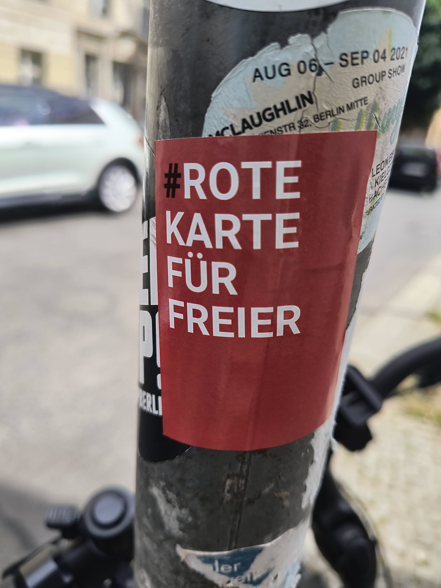 The central sticker is a rectangular, bright red sticker with bold, white sans-serif text. It has a hashtag symbol followed by the German phrase 'ROTE KARTE FÜR FREIER'. The sticker appears to be applied to a cylindrical pole, possibly a lamppost or signpost, as it is layered over other, older stickers and shows some wear around the edges, suggesting it is outdoors. The overall appearance is that of a protest or political statement sticker.