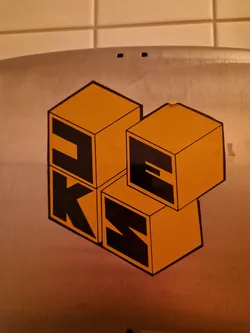 A sticker featuring a design of orange blocks forming the letters PKS.  The blocks are stacked in a three-dimensional arrangement, giving a playful and modern feel. The sticker is affixed to a metallic surface.