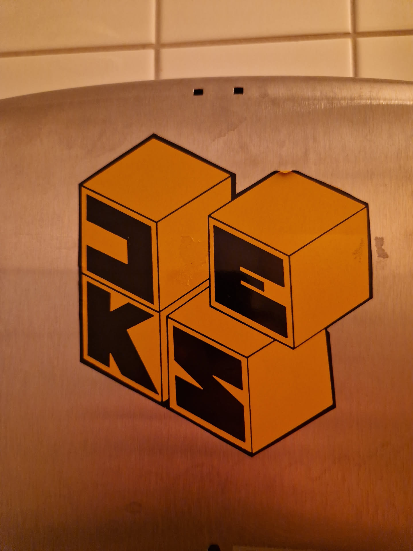 A sticker featuring a design of orange blocks forming the letters PKS.  The blocks are stacked in a three-dimensional arrangement, giving a playful and modern feel. The sticker is affixed to a metallic surface.