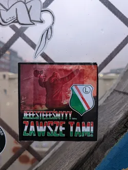 The sticker features a central image of a person in a red jacket holding a megaphone, with red smoke or flares in the background. To the right of the image is a shield-shaped crest with green and white diagonal stripes and a red stripe in the middle, containing a black circle with a white 'L' inside. Below the image, there are two lines of text. The top line is in green block letters with a white outline, and the bottom line is in red and white block letters with a green outline. The overall impression is one of energetic support, likely related to a sports team.