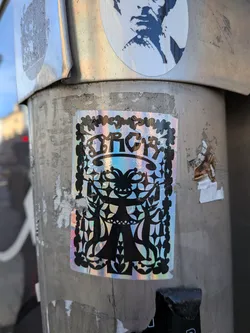 Street sticker A vertically oriented rectangular sticker with a shimmering, iridescent holographic background. The graphic consists of intricate black silhouette artwork reminiscent of traditional papercut or scherenschnitte folk art. At the top, stylized ornamental text is encased within an arched banner, surrounded by a border of hearts and leaves. Below the text stands a symmetrical, stylized jester or whimsical figure with outstretched arms, flanked by detailed geometric patterns, stars, and side silhouettes.