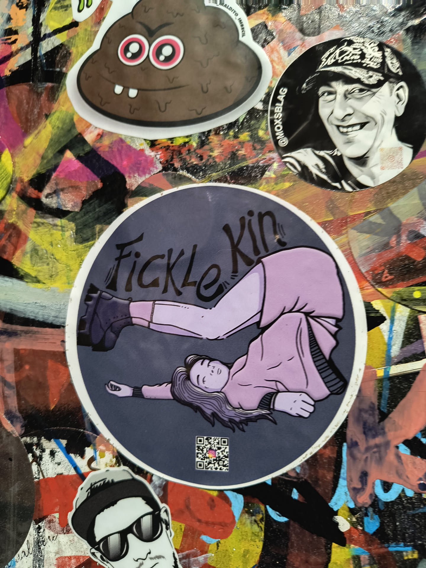 The central sticker is circular with a dark grey background. It depicts an illustration of a person lying on their back, with their legs bent and raised, one leg appearing to kick upwards. The person is wearing a light purple long-sleeved shirt and a matching purple skirt or dress. They have purple hair and purple accents on their eyes and cheeks. They are wearing dark, chunky boots. The text "Fickle Kin" is written in a stylized, dark font above the illustration. A small QR code is present in the bottom left corner of the sticker. The overall style is illustrative with bold outlines and flat colors.