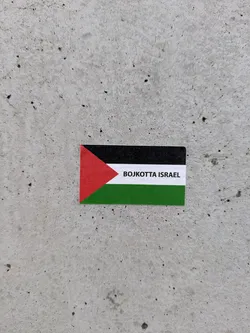 A sticker on a gray concrete surface. The sticker is in the shape of the Palestinian flag. It features a triangle of red color and a stripe of white between black and green stripes. The words "BOJKOTTA ISRAEL" are printed over the white stripe.