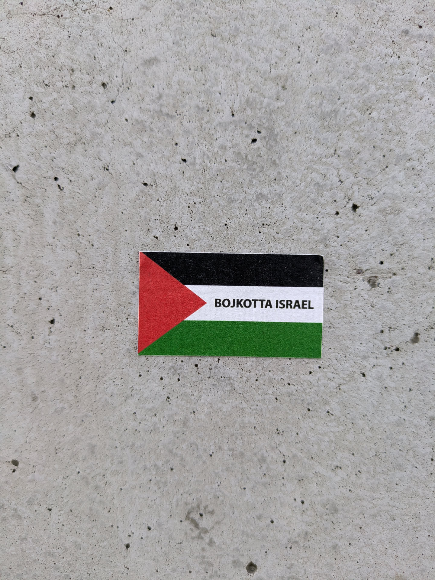 A sticker on a gray concrete surface. The sticker is in the shape of the Palestinian flag. It features a triangle of red color and a stripe of white between black and green stripes. The words "BOJKOTTA ISRAEL" are printed over the white stripe.