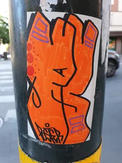 A vibrant orange sticker depicting a stylized hand. The hand is drawn with bold black outlines and features purple accents on the fingers. There's a small, illegible tag at the bottom. The sticker is affixed to a dark-colored pole.