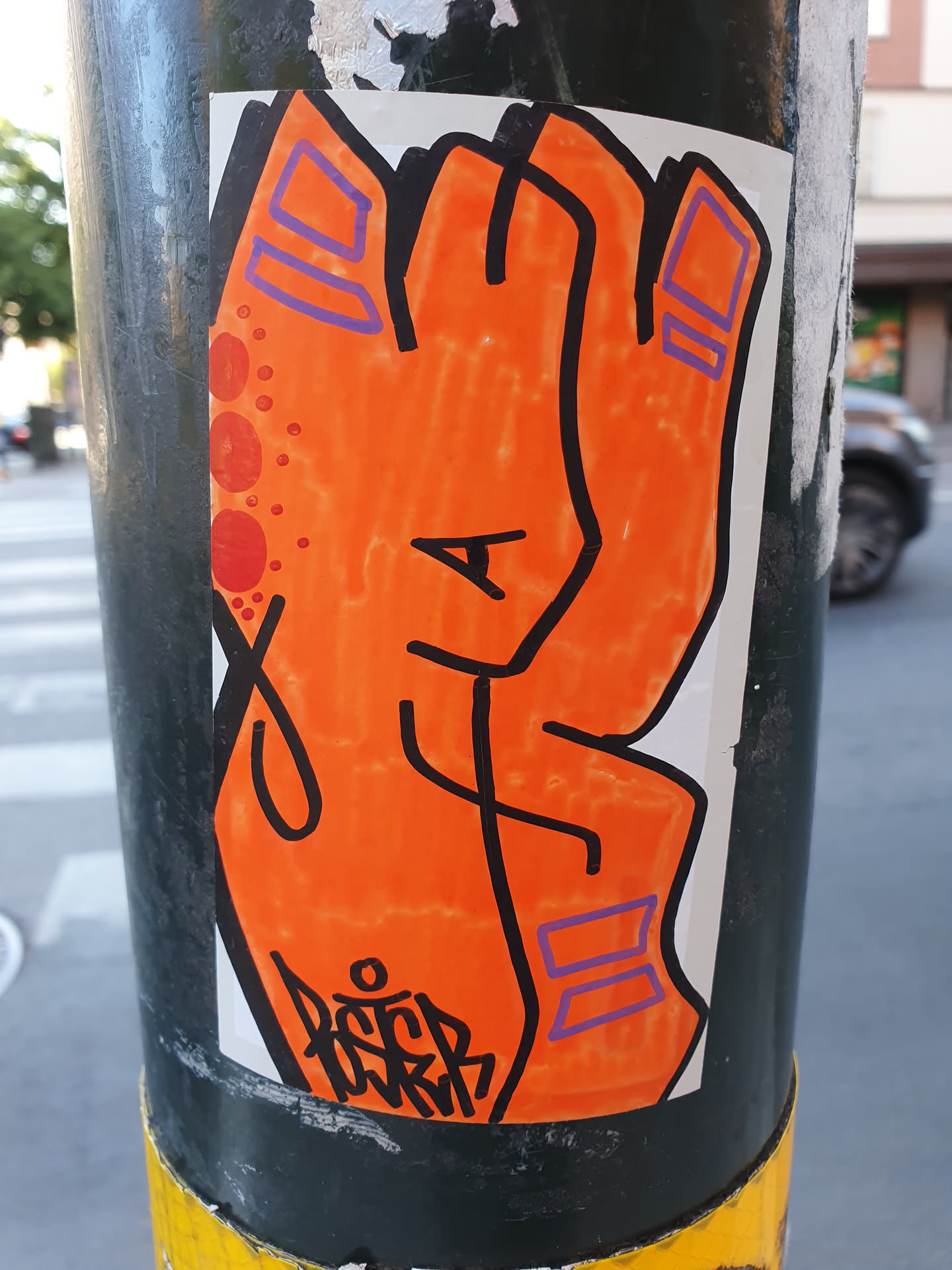 A vibrant orange sticker depicting a stylized hand. The hand is drawn with bold black outlines and features purple accents on the fingers. There's a small, illegible tag at the bottom. The sticker is affixed to a dark-colored pole.