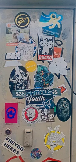 This image showcases a cluster of stickers with varied themes and styles on a weathered surface. The stickers range from artistic designs to logos and textual elements, creating a visually rich and layered composition. Some stickers are peeling or damaged, adding to the aged effect.