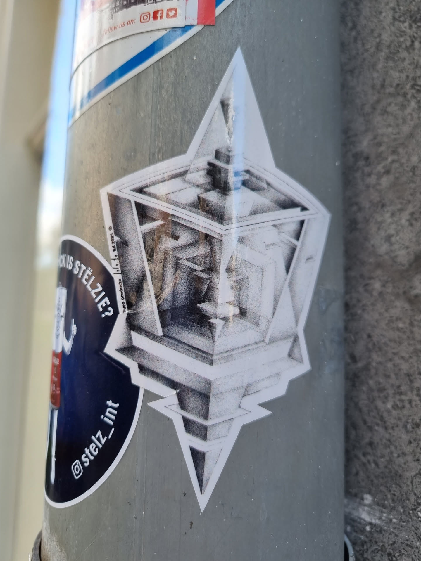 A grayscale sticker depicting a Penrose cube, a three-dimensional impossible object,  placed within a star-like geometric frame. The style is detailed and reminiscent of a pencil sketch.  A social media handle is visible in another sticker nearby.
