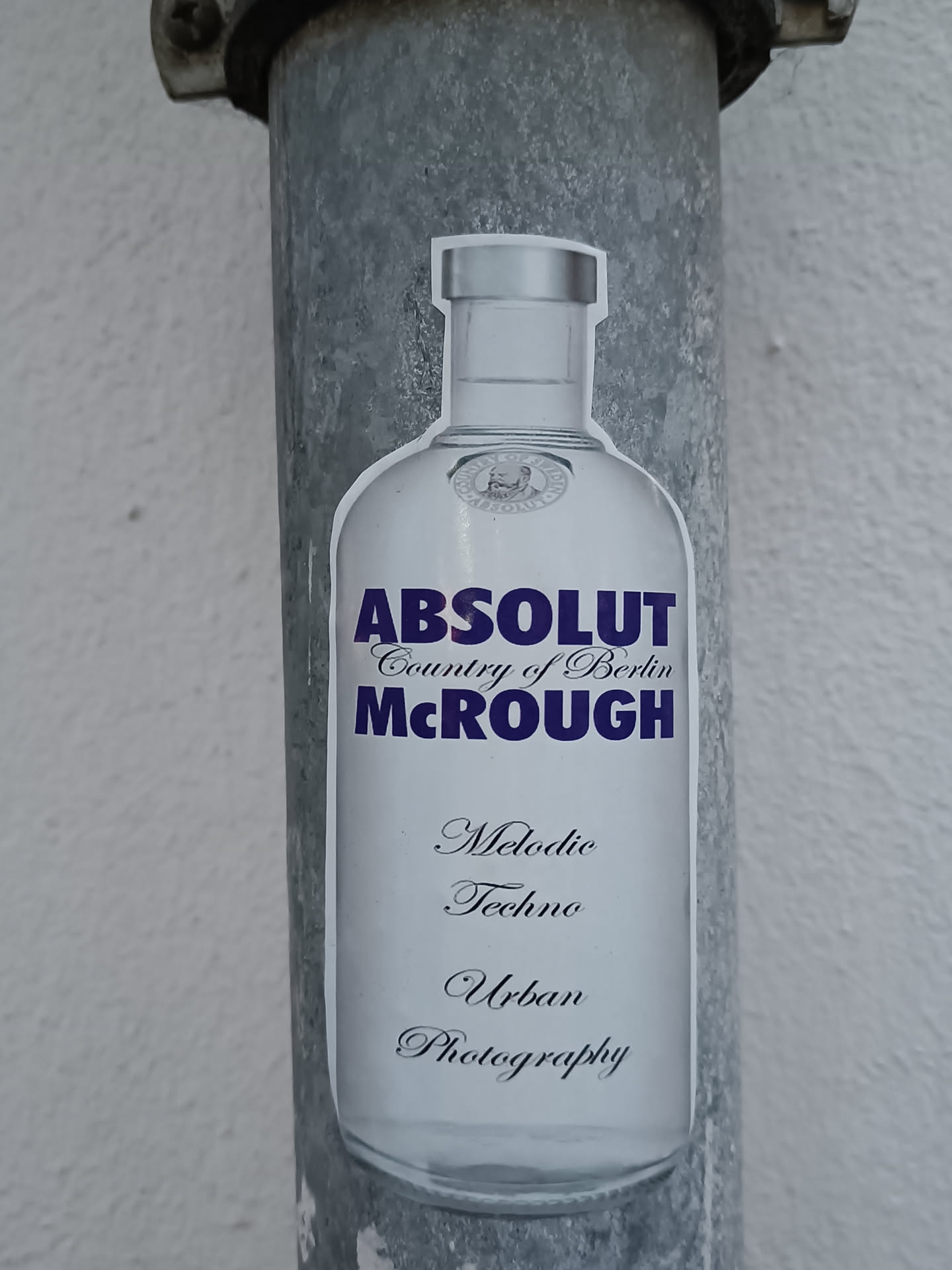 A sticker featuring an Absolut Vodka bottle with the text 'Absolut Country of Berlin McRough' and 'Melodic Techno Urban Photography' underneath. The sticker is affixed to a metallic pole.