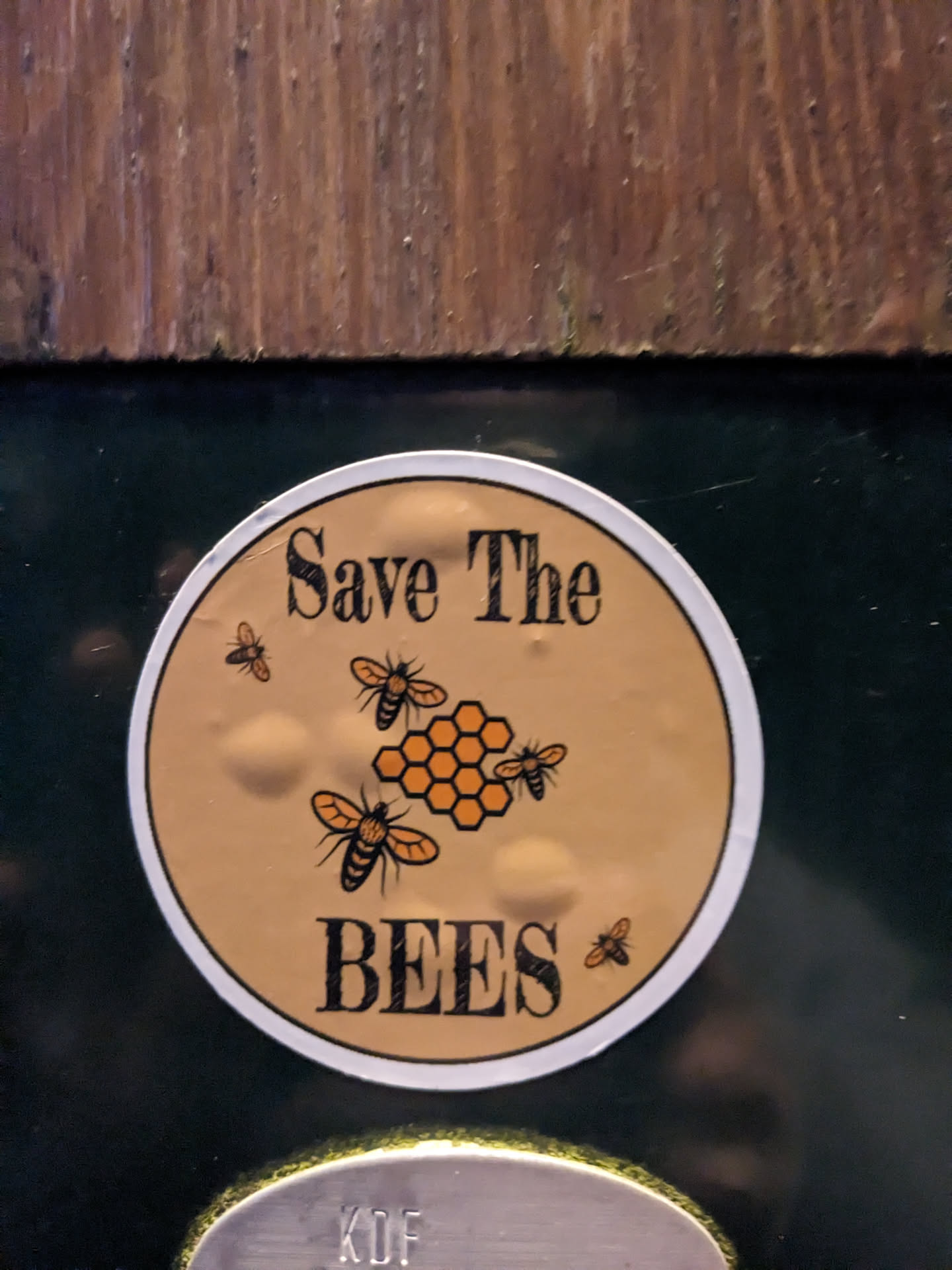 A round sticker with a beige background featuring the text "Save The Bees" and an illustration of bees around a honeycomb.  The sticker is affixed to a dark surface.