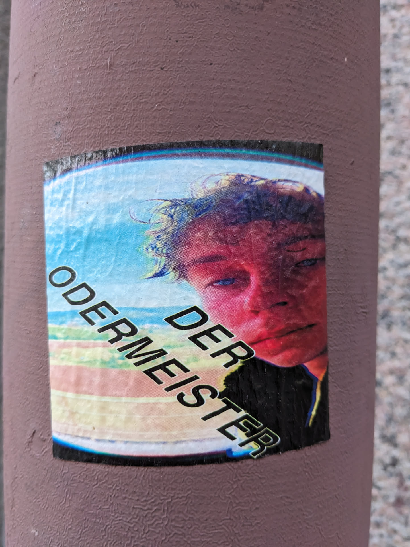 A sticker featuring a portrait of a young person with the text "DER ODERMEISTER" overlaid. The background appears to be a stylized beach or coastal scene.