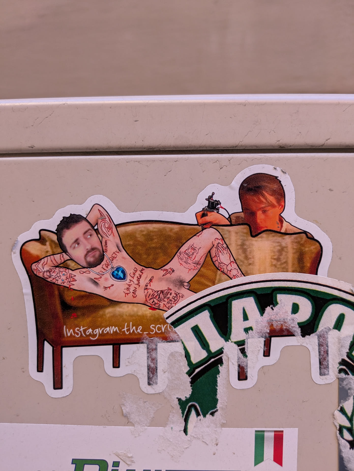 A sticker showing a naked tattooed man lying on a couch being tattooed. It seems to be a collage of an individual's face put on the tattooed model. The sticker includes social media links.