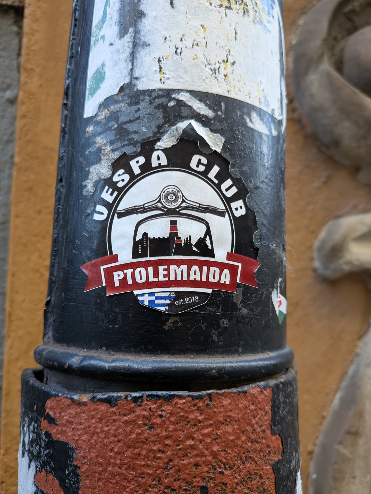 A crest-shaped emblem sticker representing the 'Vespa Club Ptolemaida'. It features a gear-toothed upper arc containing the white text 'VESPA CLUB' on a black background. At the center is a white circle showing the handlebars and round headlight of a vintage Vespa scooter framing a black silhouette of a cityscape with industrial towers/smokestacks. Across the lower part of the circle is a red ribbon banner displaying the text 'PTOLEMAIDA' in white uppercase letters. Below the banner, a small Greek flag motif and the text 'est. 2018' are visible.