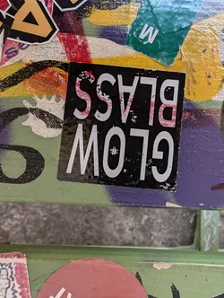 A black sticker with the words "Glow Glass" written in a stylized font. The "Glow" is written in white and the "Glass" is written in pink. The sticker is slightly worn and has some scratches.