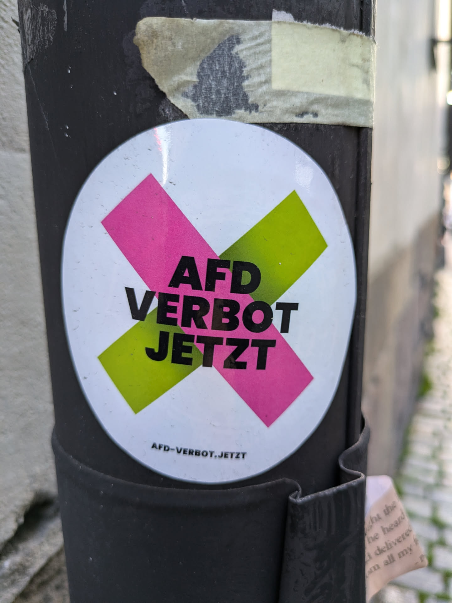 A sticker with a pink and green X over the text "AfD Verbot Jetzt", which translates to  "AfD ban now". The bottom of the sticker has a smaller text that reads "afd-verbot.jetzt"