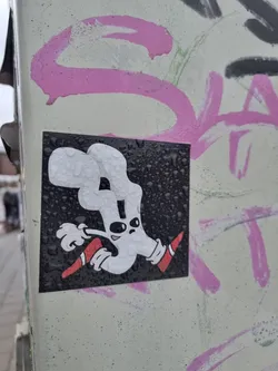 A square sticker featuring a cartoon ghost-like figure in white with red shoes, running on a black background.  The sticker is affixed to a surface with other graffiti and appears to have water droplets on it.