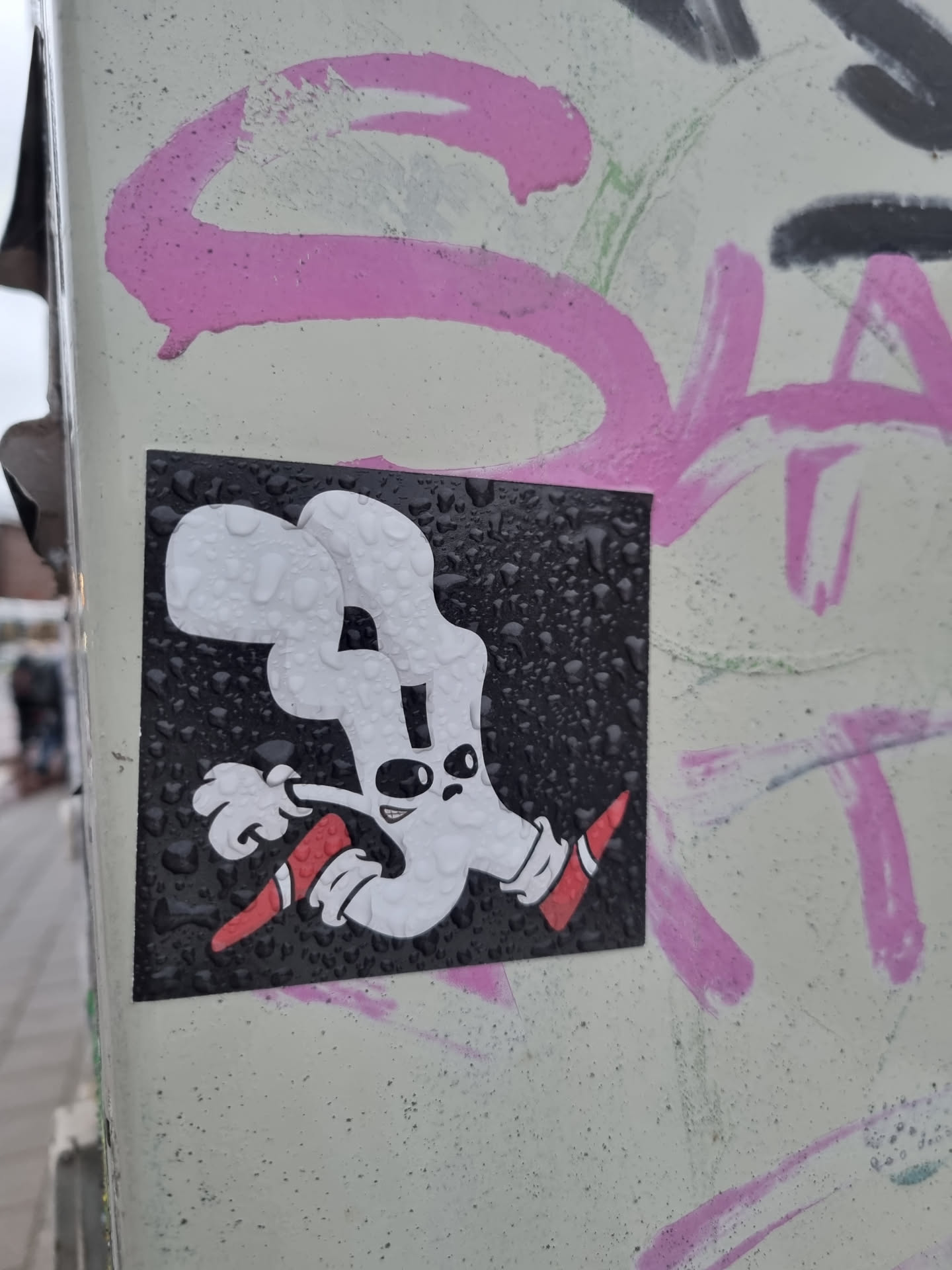 A square sticker featuring a cartoon ghost-like figure in white with red shoes, running on a black background.  The sticker is affixed to a surface with other graffiti and appears to have water droplets on it.