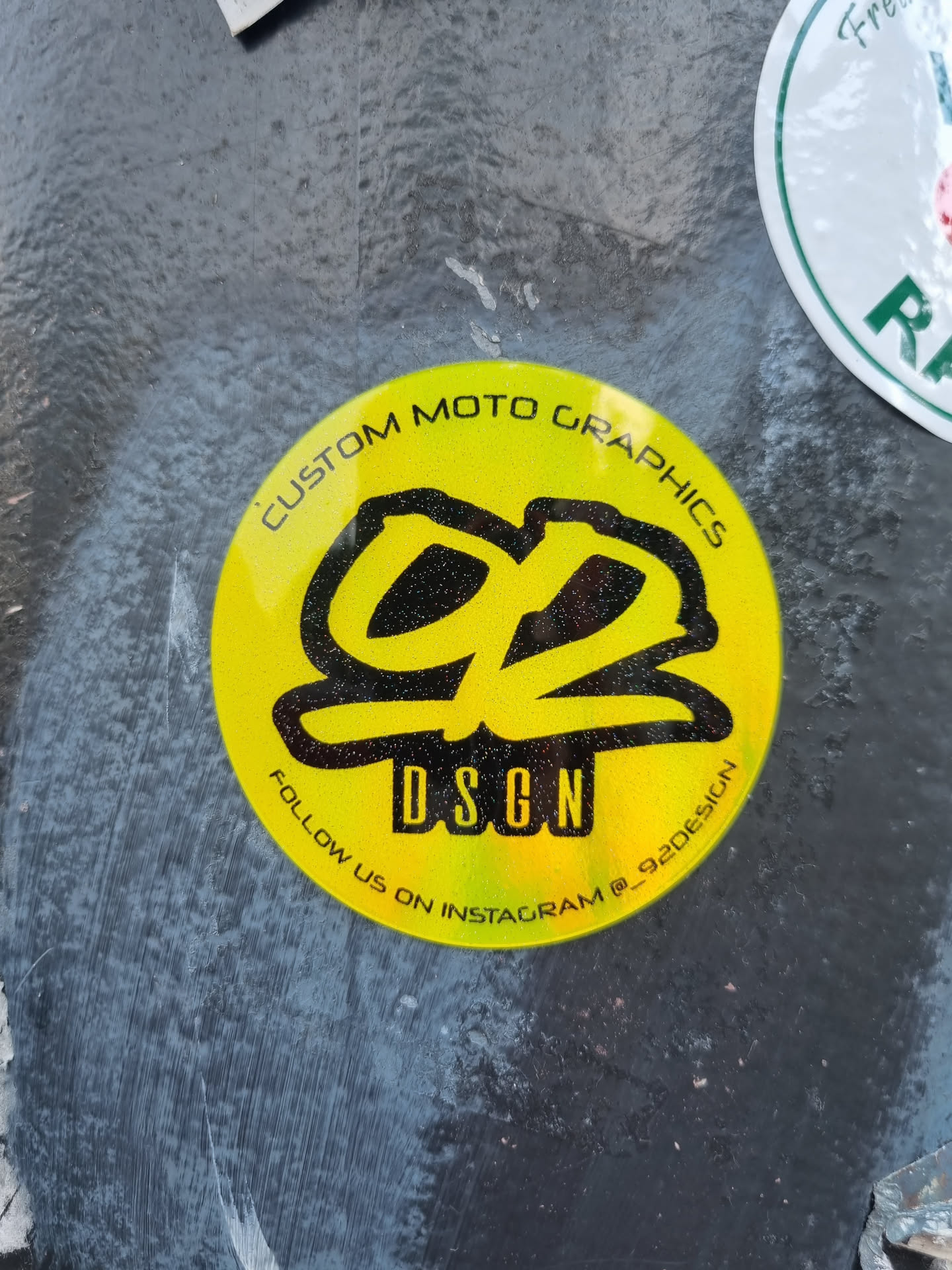 Round yellow sticker with black and glitter design. Contains the text "Custom Moto Graphics", a logo, "DSGN", and "Follow us on Instagram @92design".